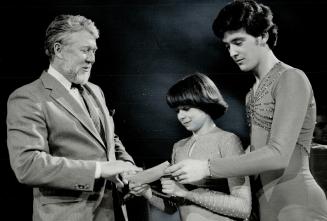 A very young girl and a young man, both in skating costumes, present a check to a bearded man i…