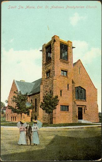 Two women in long dresses stand on a lawn beside a man in a boater hat. A large red-brick churc…