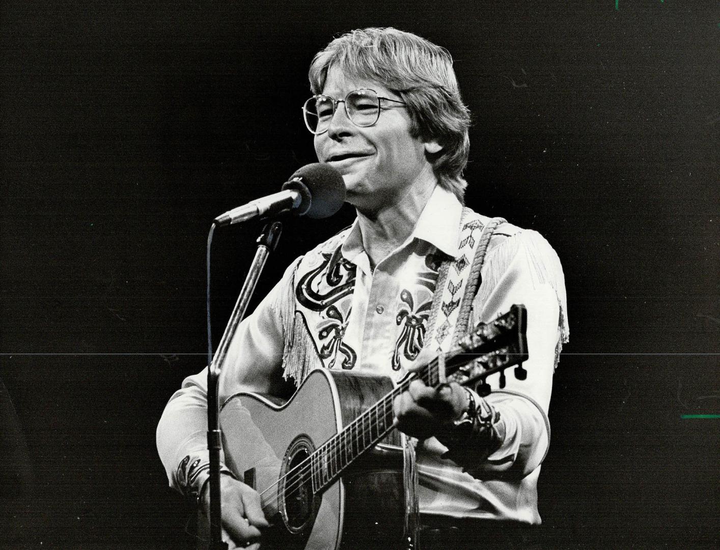 A smiling blonde man, wearing an embroidered shirt and large glasses, plays an acoustic guitar …