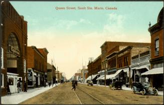 A street scene. Streetcar tracks run along the centre of an unpaved road, used by a mix of comb…