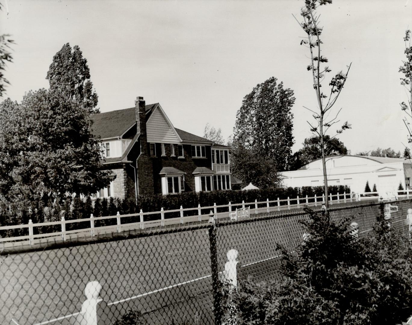 Side view of brick home seen over the top of a chain link fence, in foreground. Trees are seen …