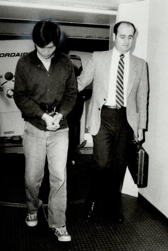 A young Asian man in handcuffs and wearing a chain on his ankles is escorted by a taller man in…