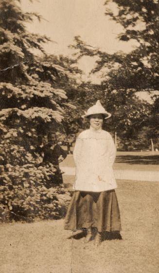 In the foreground, a woman in a light-coloured hat and shirt and long, dark dress stands smilin…