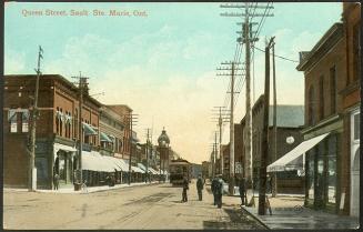 A lively street scene. A group of men stand on the unpaved roadway, a streetcar heads away from ...