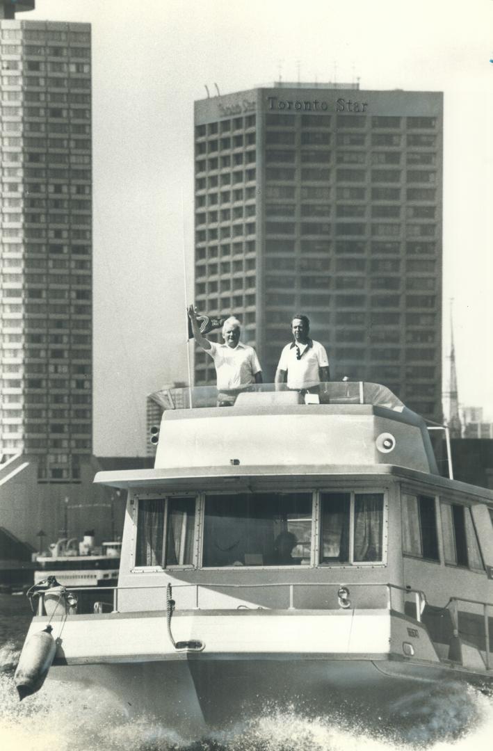 Shows a houseboat leaving Toronto harbour with the Toronto Star building in the background. Two…