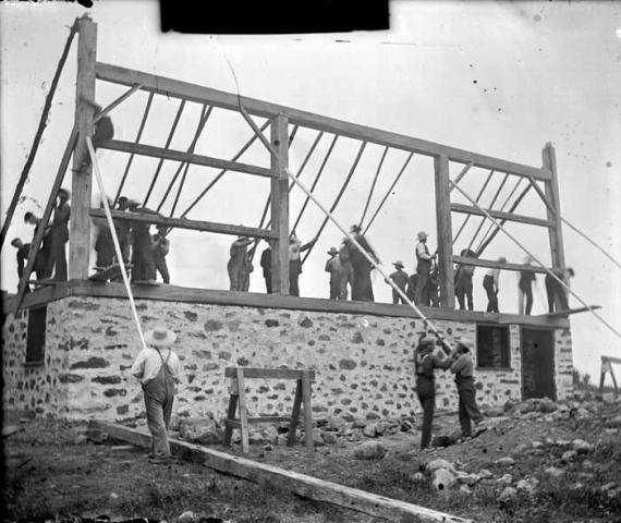 Shows a group of 25 to 30 men, in suspenders and hats, raising a frame for a wall onto a stone …