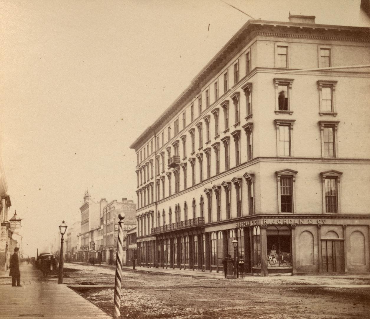 Corner view of a 1860s-era 5-storey hotel. There is a barber pole and a gas streetlight across …