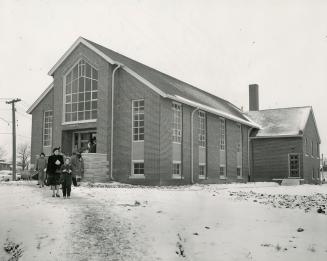 Small crowd seen exiting gable-roofed church with lattice window above entrance.