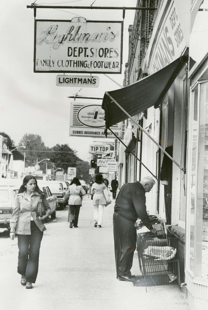 Shows a sidewalk scene. Women in 1970s attire walk down the sidewalks. An older man looks throu ...