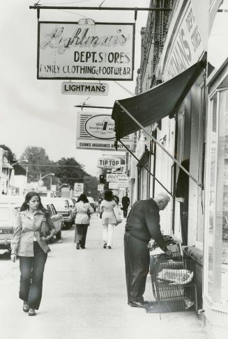 Shows a sidewalk scene. Women in 1970s attire walk down the sidewalks. An older man looks throu…