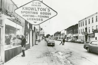 A man in a plaid jacket, carrying a metal lunch pail, looks into the window of a shop. A sign r…