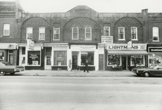 Shows ornate brick storefronts. A couple and a dog walk along the sidewalk. 1970s-era cars are …