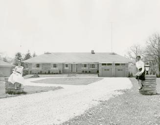 Driveway leading up to bungalow with two-car garage. Two women, left and right of driveway, sit…