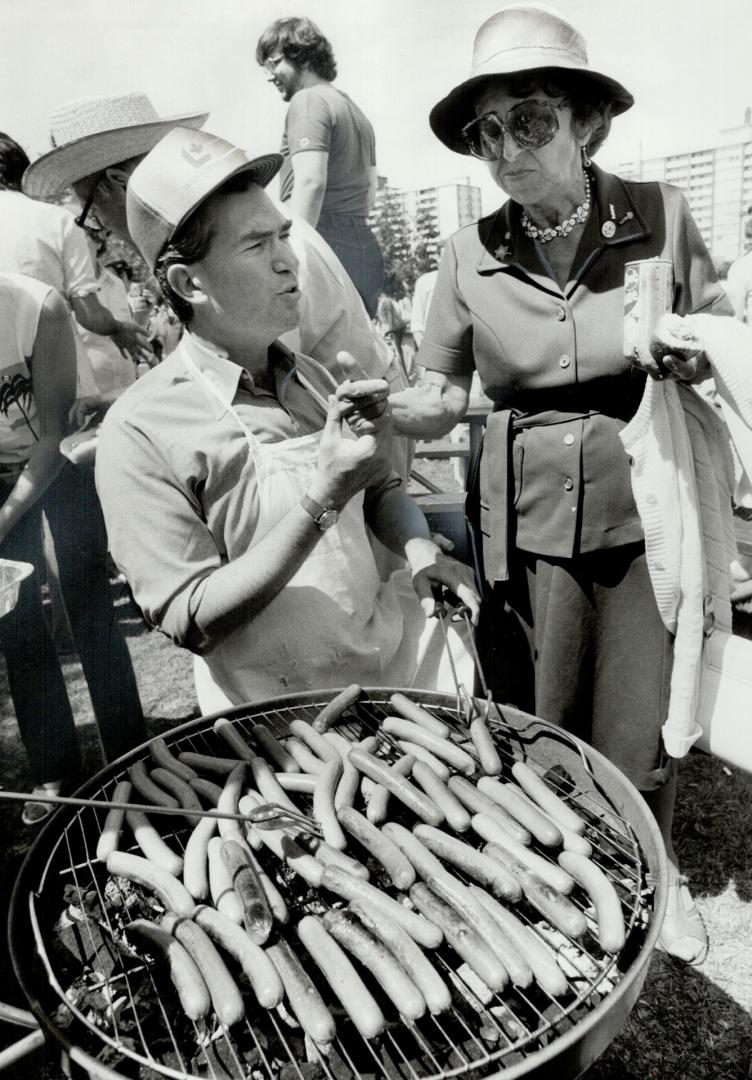 A man in a Liberal baseball cap stands in front of a barbecue grill full of hotdogs, while he s…