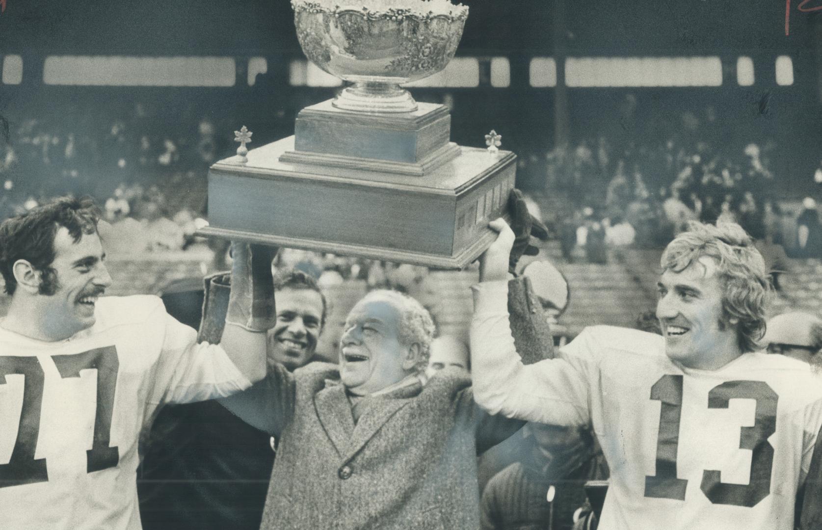 Winning captains of St. Mary's Huskies, Fraser MacDonald (77) and Ken Clark (13) get assist from Governor-Gneral Roland Michener in holding aloft Vani(...)