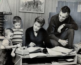 Mrs. Merrill works on accounts while family watches. Husband Frank watches bookkeeping but isn't worried. Brad, 1, and Frankie, 3, check too