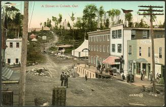 Colorized photograph of people walking on a dirt street of a newly built town with wooden build…