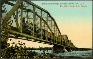 A long steam train on a trestle spanning a large river, with bushes in the foreground. 