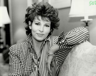 A woman with short curly hair smiles at the camera while sitting on a chesterfield in a living  ...