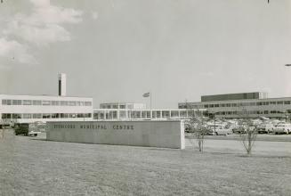 Light brick, two and three-storey adjoined buildings stand beyond a parking lot full of cars. A…