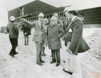 Several men in suits and hardhats stand in front of multi-level, flat-faced building with row o…