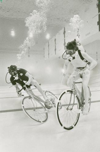 Two barefoot men ride bikes underwater in a white tiled pool; they wear scuba tanks strapped to…