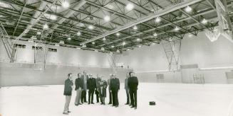 Small group of men and one woman stand in brightly lit empty gymnasium, looking in direction of…