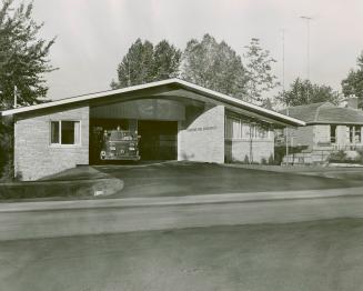 Brick bungalow with wide driveway leading to two-car garage. Fire truck protrudes from left sid…