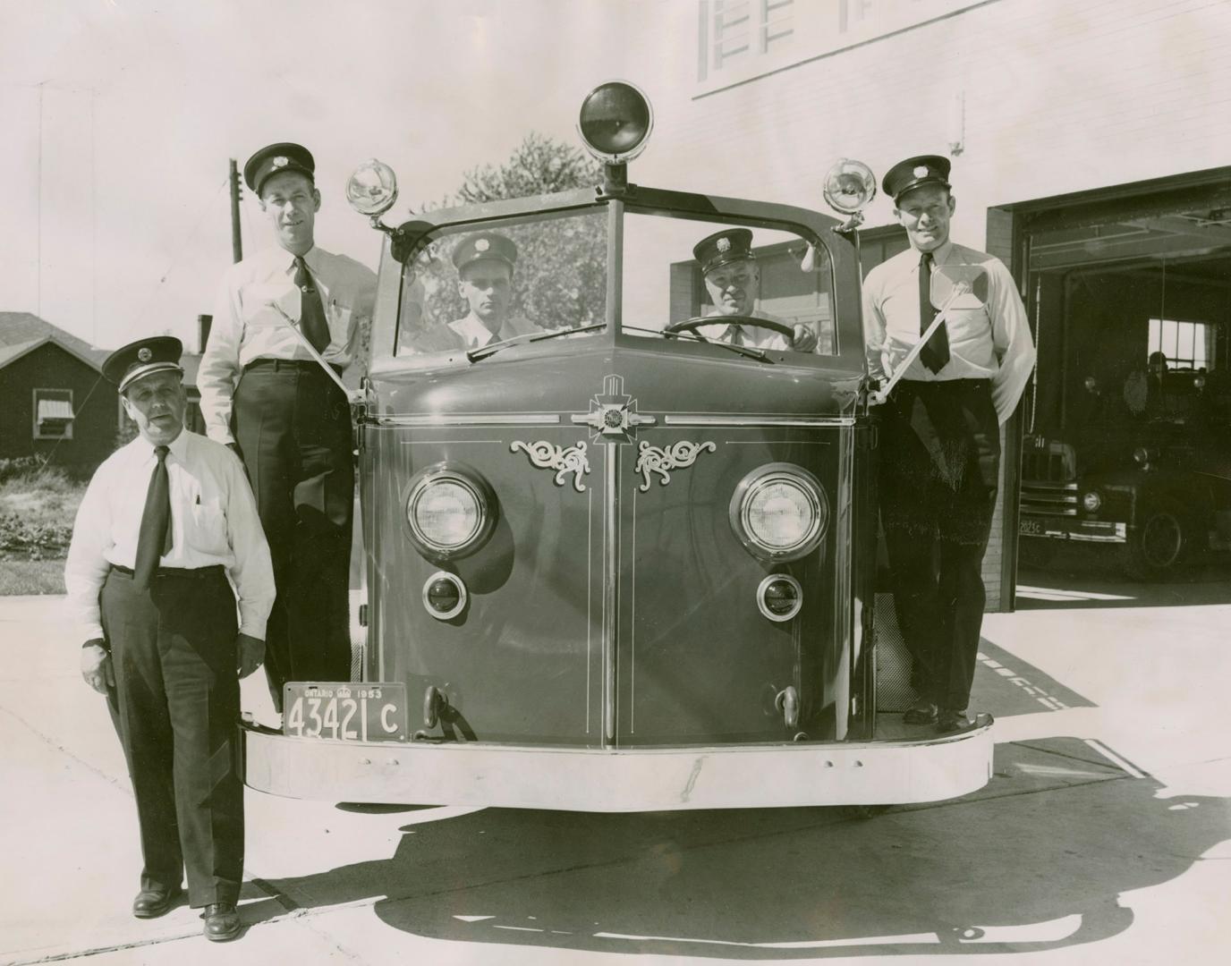 Four firemen in white shirts, black pants, ties and peaked officer caps, pose with fire engine.…