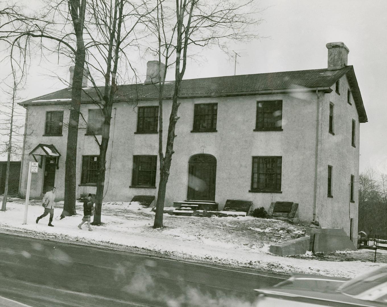 Two-storey building with dark framed windows and doors, shingled roof, and three prominent ston…