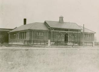 One-storey brick building with arched windows either side of arched entrance. Enclosed school b…