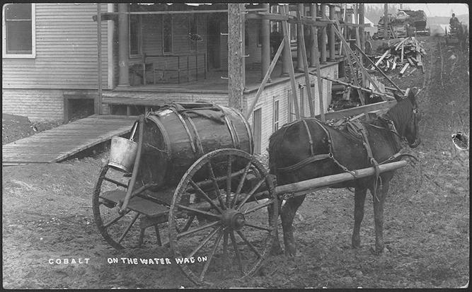 Black and white photograph of a horse pulling a wagon with barrel in front of a building with a…