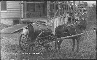 Black and white photograph of a horse pulling a wagon with barrel in front of a building with a…