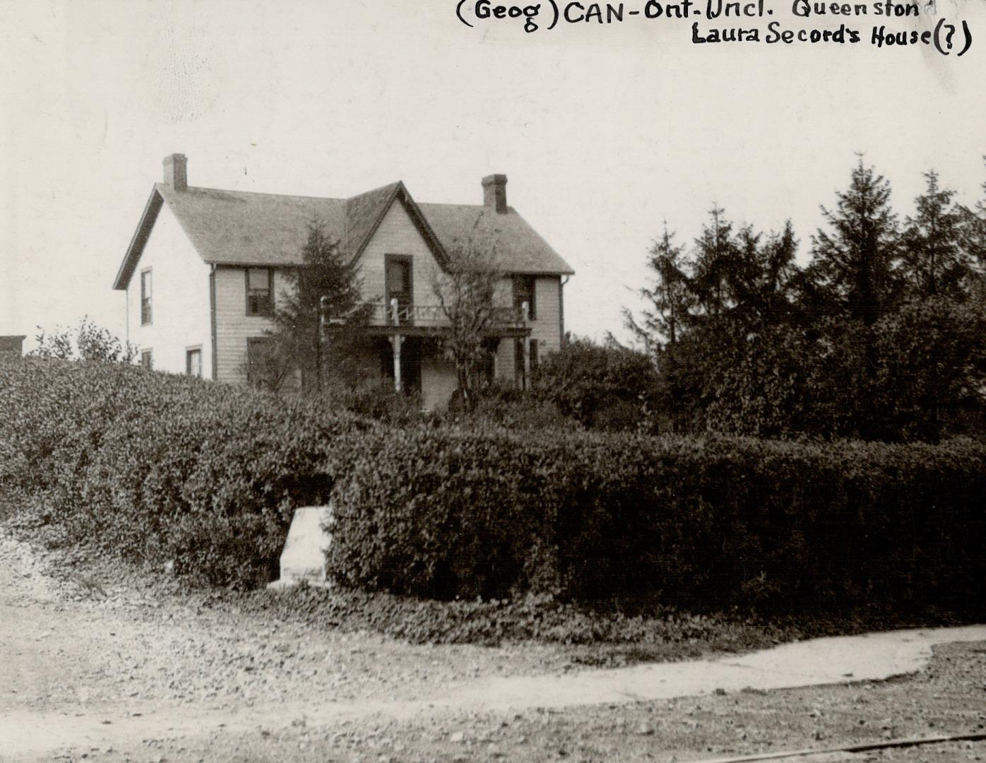 A photo of a two-storey wooden cottage with a steeply-pitched roof, two chimneys, surrounded by…