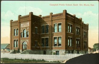 A blocky two-storey red-brick building surrounded by a white picket fence. Two men with flat ca…