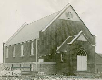 Brick church with stone foundation, steeply pitched roof and covered entrance. Wood planks pile…