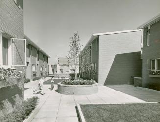 Paved pathway bordered by blocks of lawn and shrubs runs between two rows of brick townhouses. …