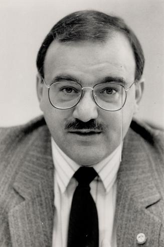 A close-up of a mustached man, with a pin-striped suit and round glasses, looking into the came…