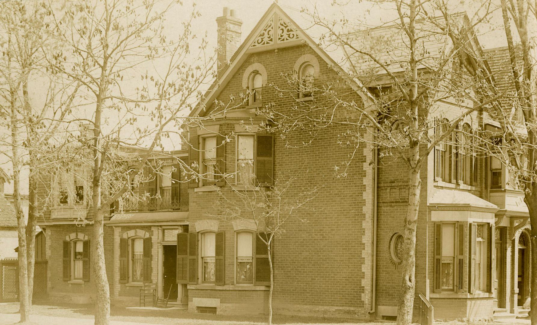 Side view of a large, ell-shaped home with bow windows and wooden lace on the edge of the roof.…