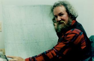 A man with long grey hair wearing a red jack shirt holds a map while looking at the camera.