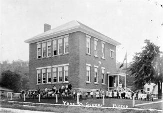 Shows a two-storey brick schoolhouse with sash windows and small verandah on the side. School c…