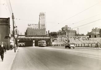 Image shows a street view with a few cars on it and some pedestrians on both sides and a railwa…