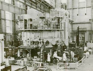 Several men in white coats work at the base of a large piece of industrial equipment.