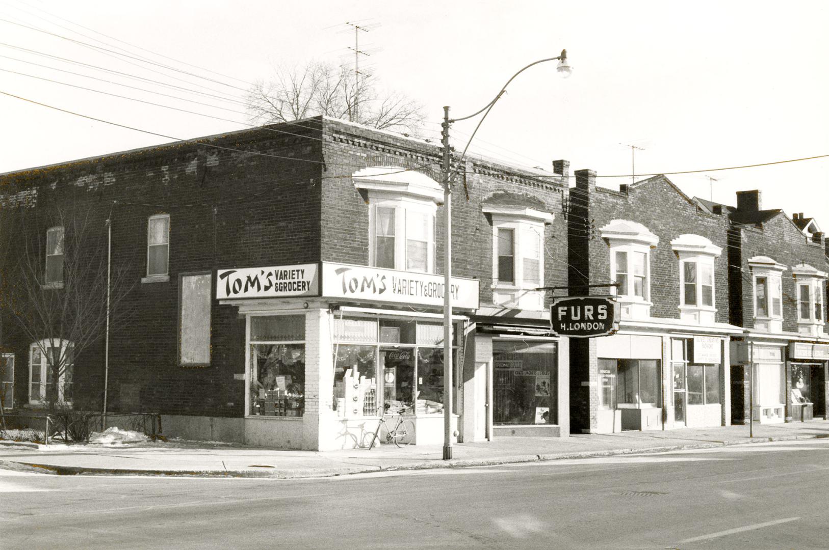 A corner shot of three duplex-style storefronts, the left-most of which is Tom's Variety Grocer…