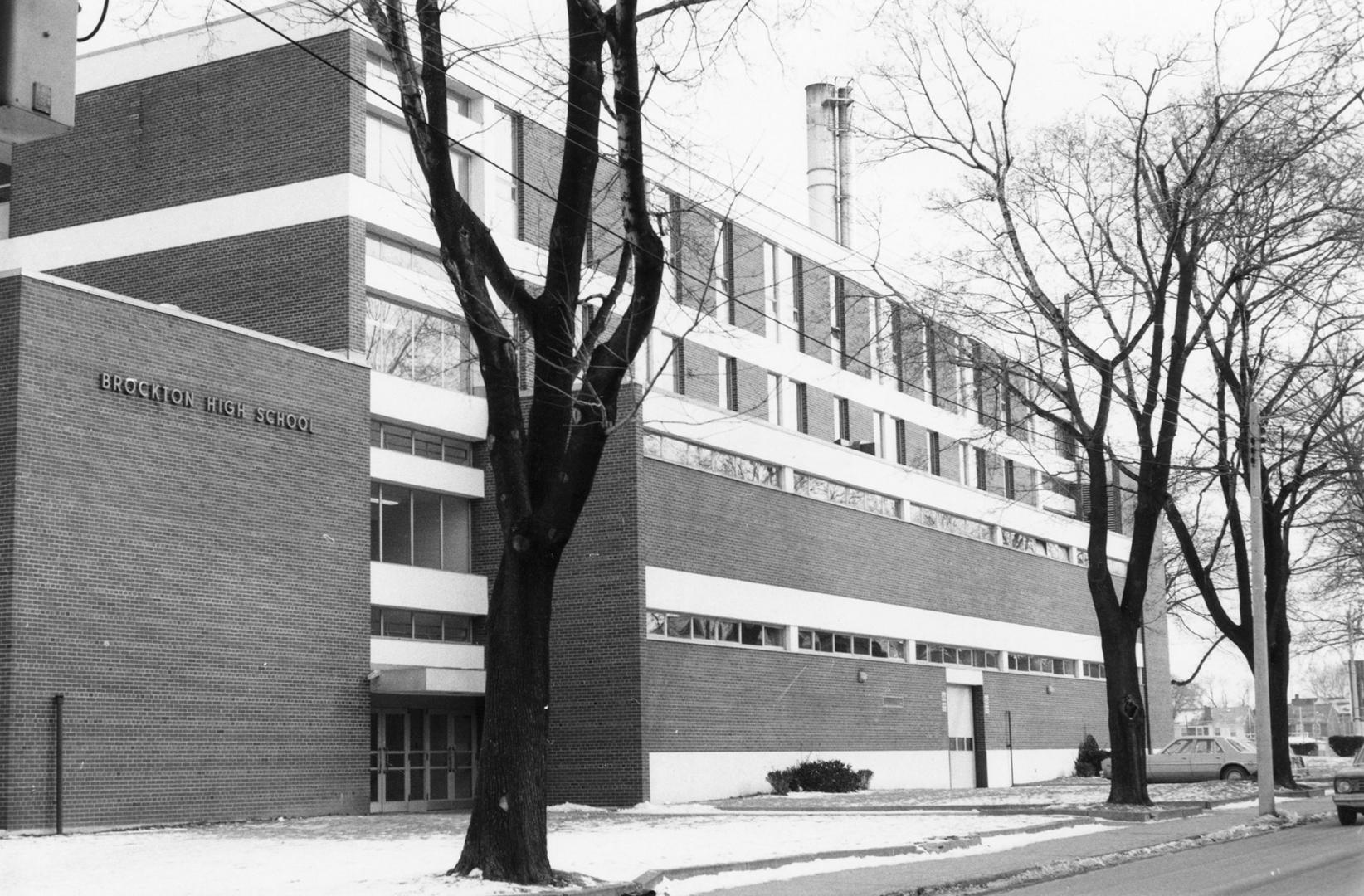 Shows a three-storey brick building with leafless trees in the snowy lawn in front of the entry…