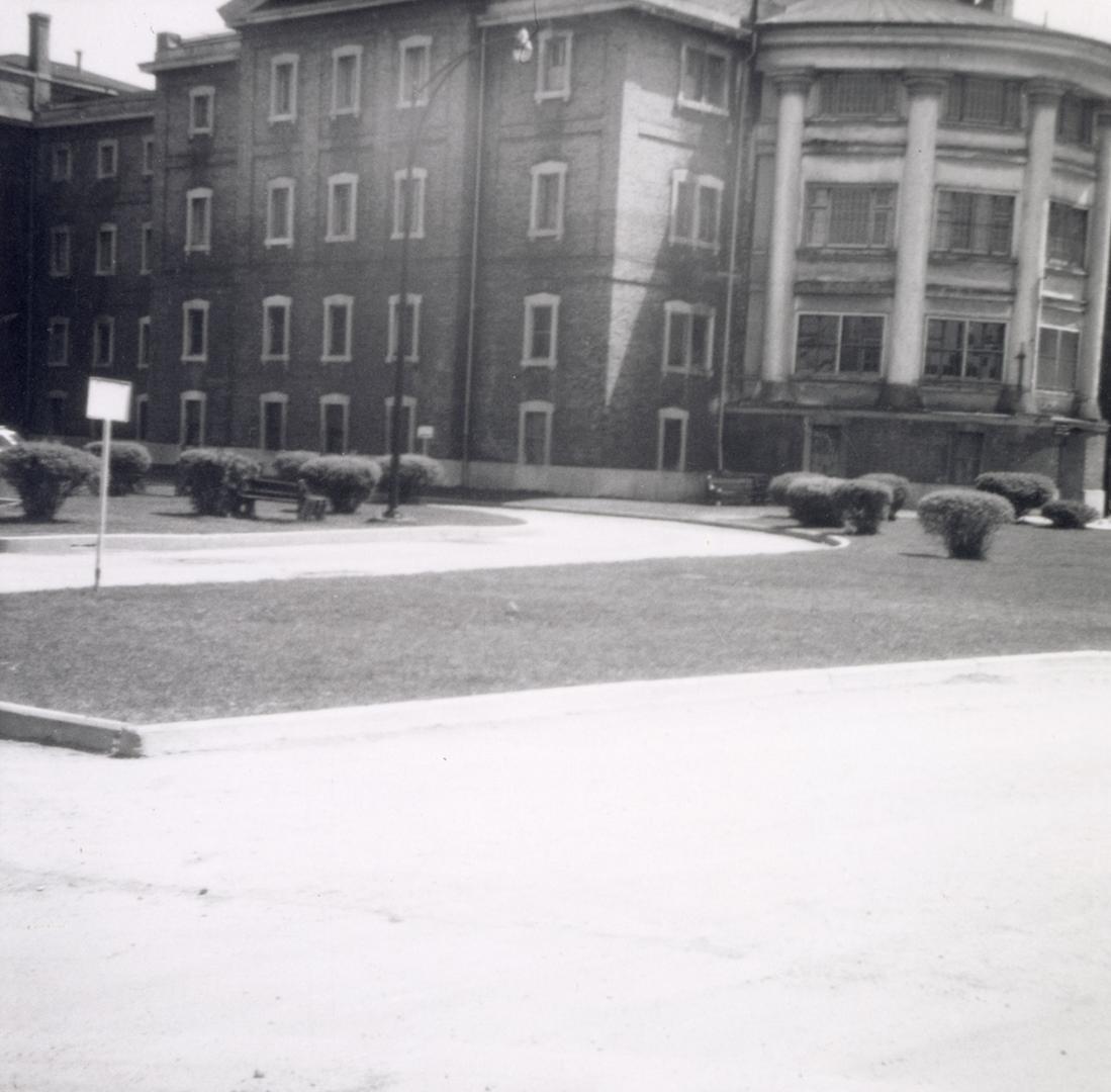 Shows a large brick four-storey building, with a rounded section ornamented by colonnades on th…