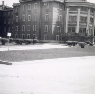 Shows a large brick four-storey building, with a rounded section ornamented by colonnades on th…