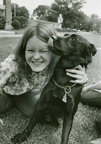 A girl crouches in the grass near a small black dog. She hugs the dog while it licks her forehe ...