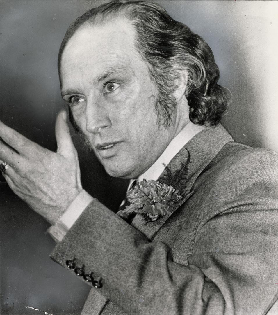 A close-up of a man with longish hair and side whiskers, wearing a carnation, speaking intently…