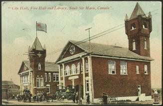 Two red-brick, two-storey buildings with towers. Several people stand in front, near horses and…